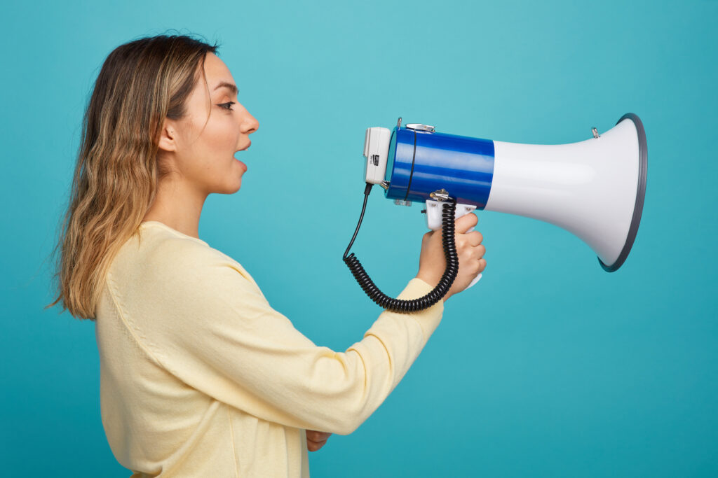 A woman in a yellow shirt speaking into a blue and white megaphone against a turquoise background.